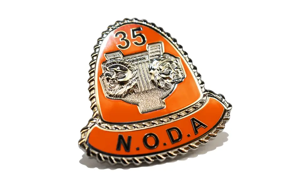 Die stamped, silver plated NODA Badge with orange enamel inlay.