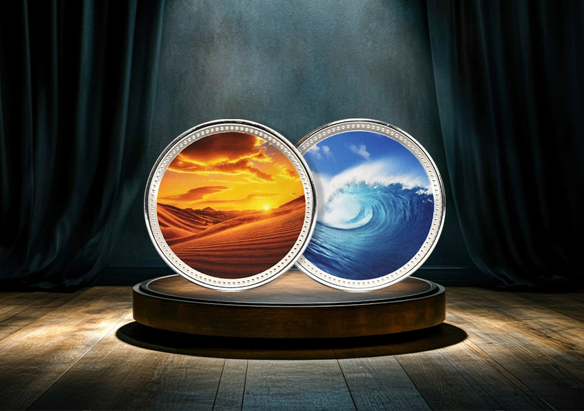 Two silver-plated custom collector coins displayed on a stage plinth, desert dunes sunset and ocean wave designs, ECO UV colour print
